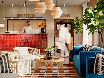El vestíbulo de un hotel con un recepcionista tras un mostrador de azulejos rojos brillantes, una zona de estar con sofá azul y sillas de mimbre, y un huésped difuminado caminando.
