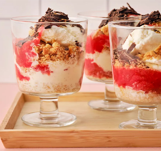Three glass cups filled with layered parfaits containing cream, crushed biscuits, red fruit sauce, and shaved chocolate on a wooden tray.