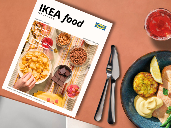 Front cover of "IKEA food MAGAZINE" placed on a terracotta-colored table next to a plate with a meal, cutlery, and a glass of pink drink.