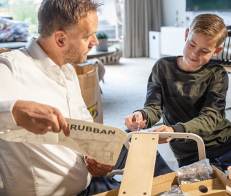 Man reads GRUBBAN furniture manual while a boy assembles the frame at home.
