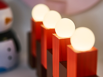 Red light decoration with illuminated white globes, displayed on an indoor surface. A blurred snowman decoration appears in the background, creating a cozy festive atmosphere.