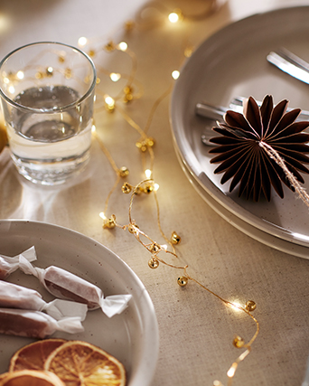 Festive table setting with neutral-toned plates, a glass tumbler, wrapped sweets, dried orange slices and a warm string of lights. A folded paper ornament adds an elegant seasonal touch.