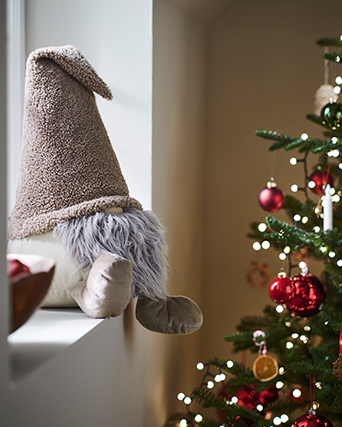 Soft fabric gnome decoration in beige and grey tones, displayed beside a Christmas tree decorated with red baubles and warm lights.