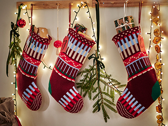 Knitted Christmas stockings in red and white tones hanging on a wall decorated with fir branches, lights and festive ornaments.