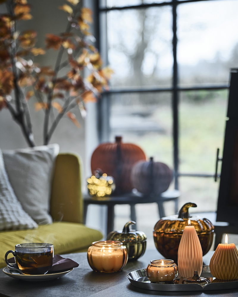 A cosy corner next to a window has a coffee table with lit HÖSTAGILLE scented candles, pumpkin-shaped pillar candles and mug.