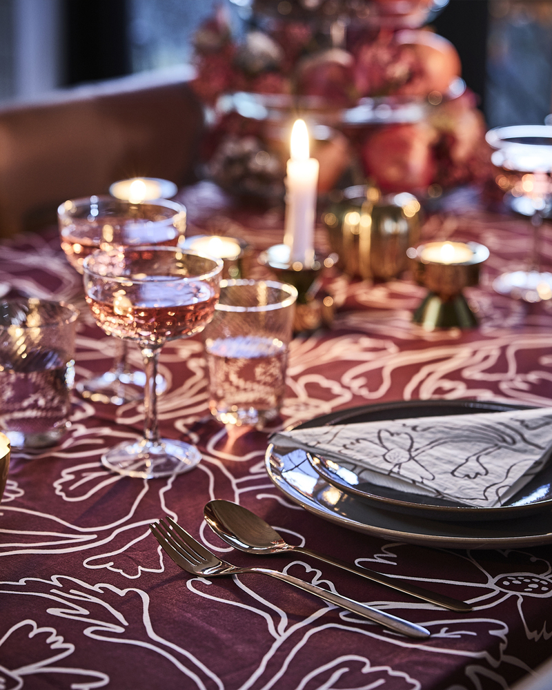 A pink and red table dining table is set with lit candles, a red patterned HÖSTAGILLE tablecloth, paper napkins and coupes.