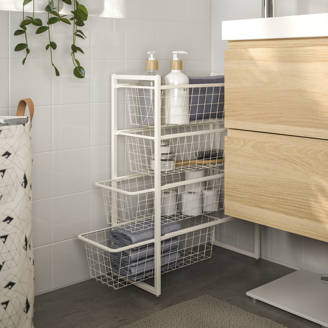 Carrito organizador extraíble de malla metálica blanca con artículos de baño, toallas y papel higiénico junto a un mueble de lavabo de madera.