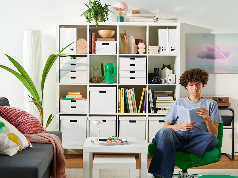 Young man reading a book in a green armchair next to a white cube shelving unit.
