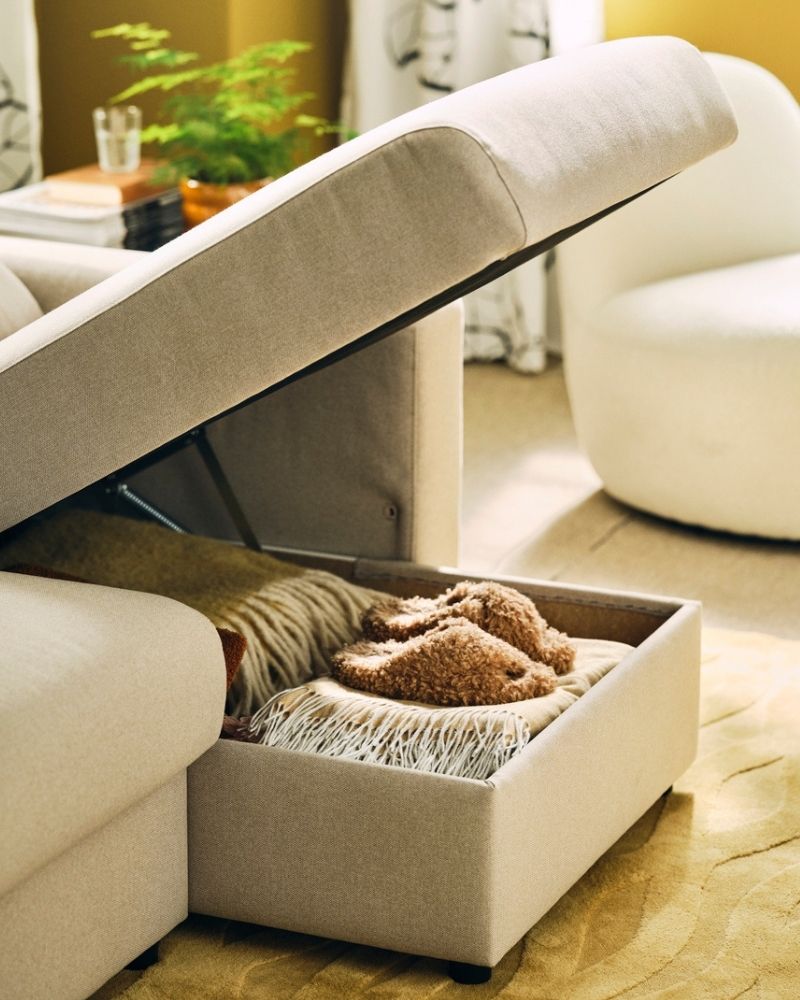 A living room with a light-beige MALMKÖPING three-seat sofa with its chaise longue open, revealing stored textiles