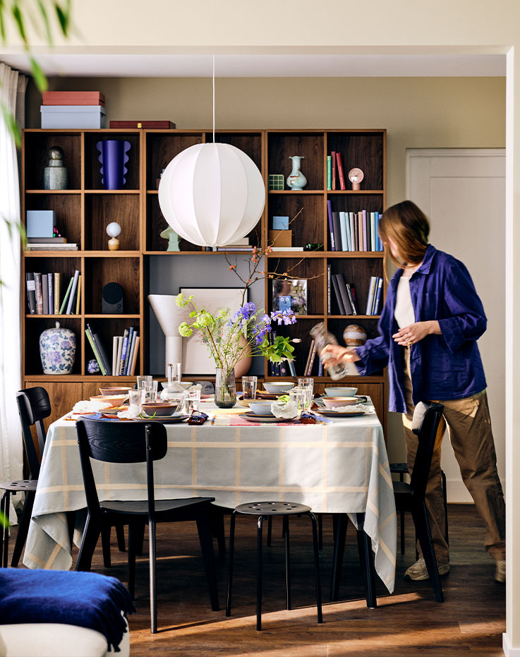 Dining room with a LISABO table and chairs, and a person coming over to set the table for dinner before the day ends.