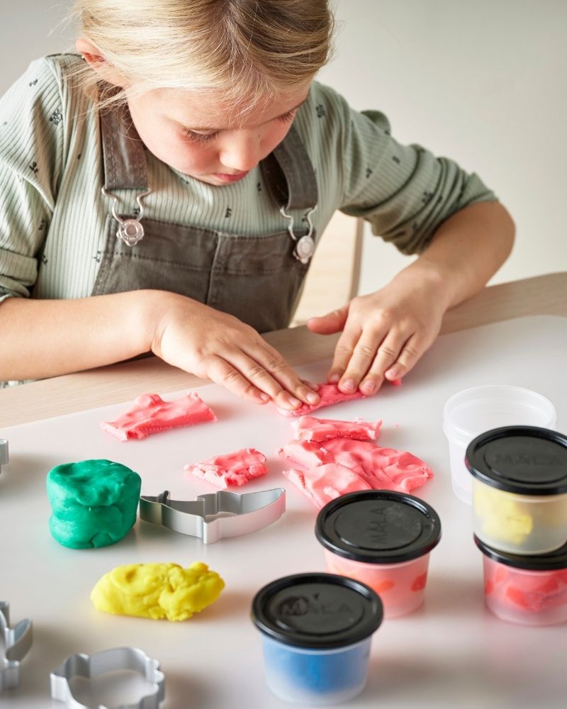 A young girl is sitting at a table, sculpting figurines with colourful MÅLA modeling dough.