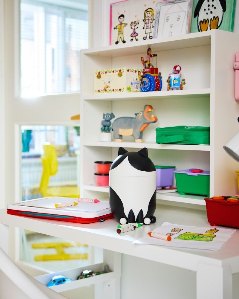 In a bright children's room, a cat-shaped GREJSIMOJS storage with lid is on a desk with colourful boxes and other items.
