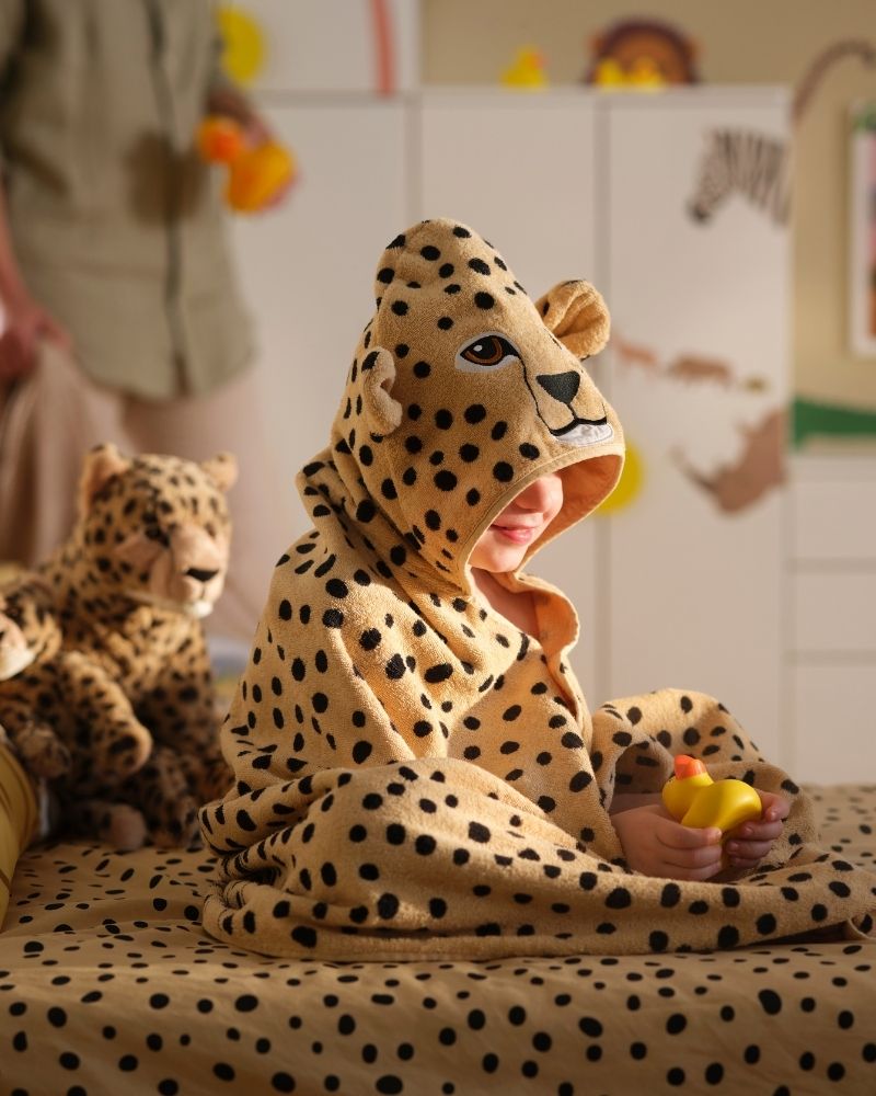 A child in a SANDLÖPARE towel with hood with cheetah pattern sits on a bed surrounded by soft toy cheetahs and a rubber duck.