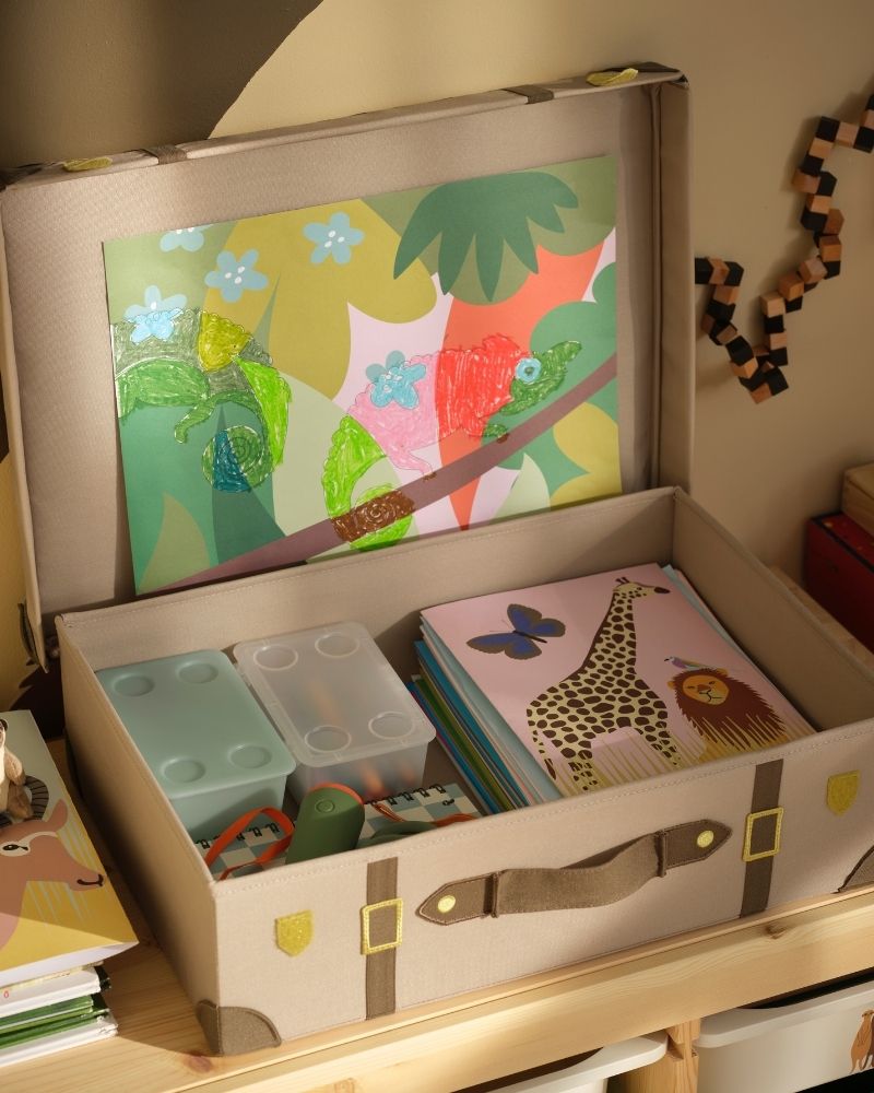 A light brown SANDLÖPARE storage trunk is opened on a wooden bench. Inside the trunk, art supplies are neatly stacked.