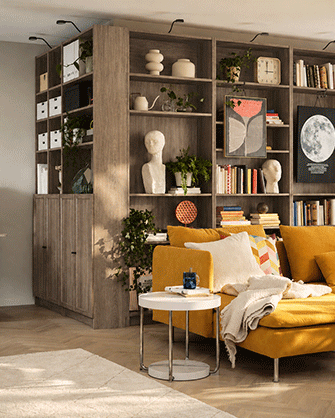 Salón moderno dividido por estanterías altas de madera oscura, con sofá amarillo, mesa auxiliar blanca y zona de trabajo al fondo.