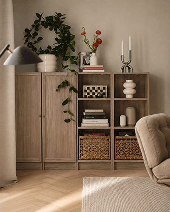 Librería de madera oscura llena de libros, láminas y cestas de fibra natural detrás de un sofá amarillo con cojines.