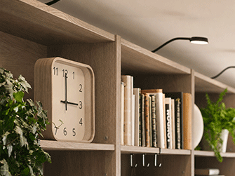 Reloj cuadrado de madera clara en estantería de nogal oscuro junto a libros, plantas verdes y foco superior.