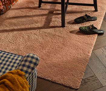 A STOENSE rug in pale pink is on a living room floor. It has a black coffee table on top and a chequered armchair next to it.