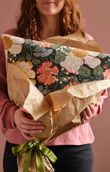 A person in a pink room holds a cushion with a floral ARENBOK cushion cover wrapped in brown paper with a green ribbon.