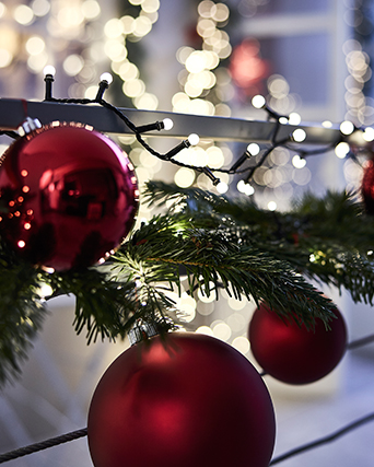A lit outdoor STRÅLA lighting chain, a green VINTERFINT artificial garland and large red tree decorations on a balcony rail.