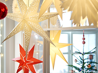 A group of white and red glowing STRÅLA star lampshades hanging from a ceiling with a large red VINTERFINT tree decoration.