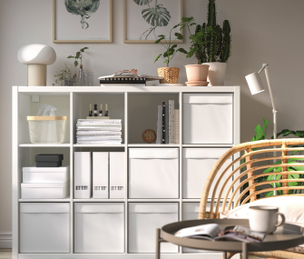 Estantería modular blanca organizada con cajas, plantas de interior y un sillón de mimbre.