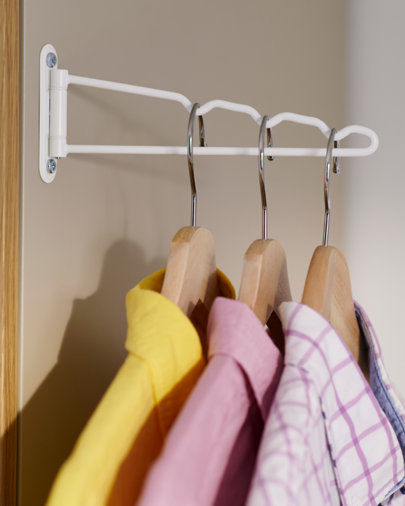 White pull-out closet rod holding wooden hangers with colorful shirts.