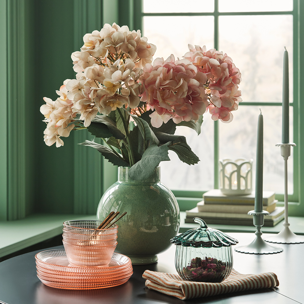 A bouquet of SMYCKA artificial flower in light pink and beige stand in a green vase on a table with glass plates and bowls.