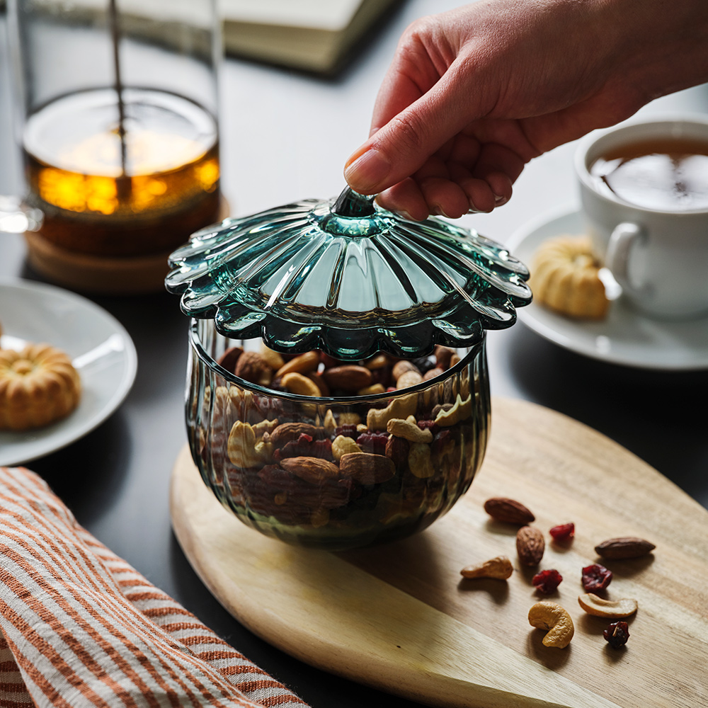 A SNÖSISKA bowl with lid in dark grey-green glass stands on a wooden cutting board. The bowl has nuts in it.