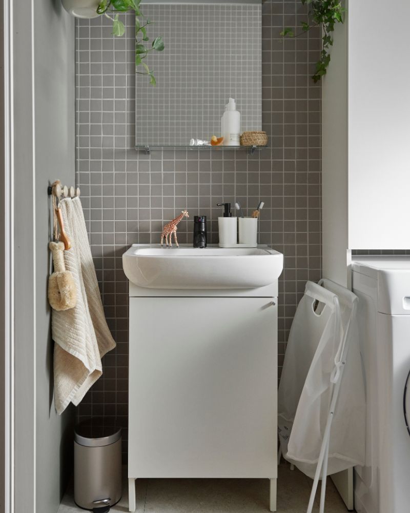 Un baño pequeño con azulejos grises, lavabo blanco, toallas colgadas y cesto de ropa. Ambiente funcional y compacto.