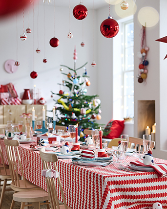 Comedor decorado para Navidad, con una mesa preparada con vajilla y textiles en rojo y blanco, adornos navideños colgantes y un árbol de Navidad al fondo.
