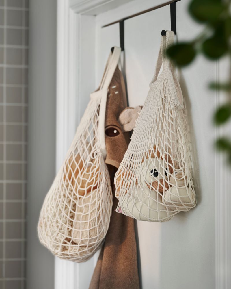 Dos bolsas de malla blancas cuelgan de ganchos, con juguetes de peluche y una toalla marrón. Ambiente infantil y ordenado.