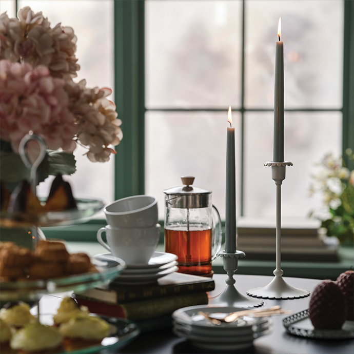 Table set with French press, cups, pastries, and tall lit candles by the window.