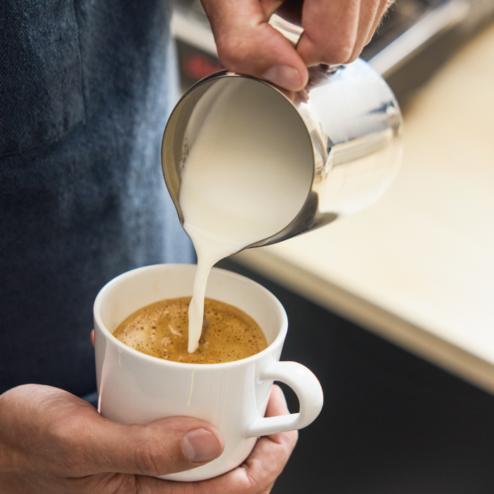 Una persona vertiendo leche espumosa de una jarra metálica para preparar café con leche en una taza blanca.