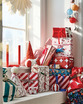 Wrapped Christmas gifts stacked beside a window decorated with lights and holiday ornaments.