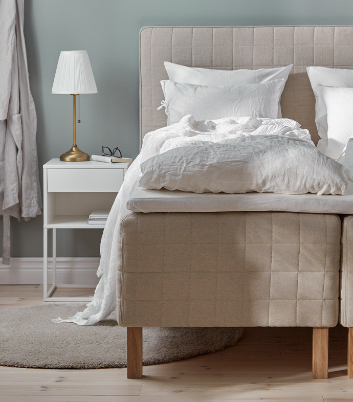 Beige upholstered bed with crisp white linen bedding beside a white nightstand and a brass table lamp with a pleated shade.