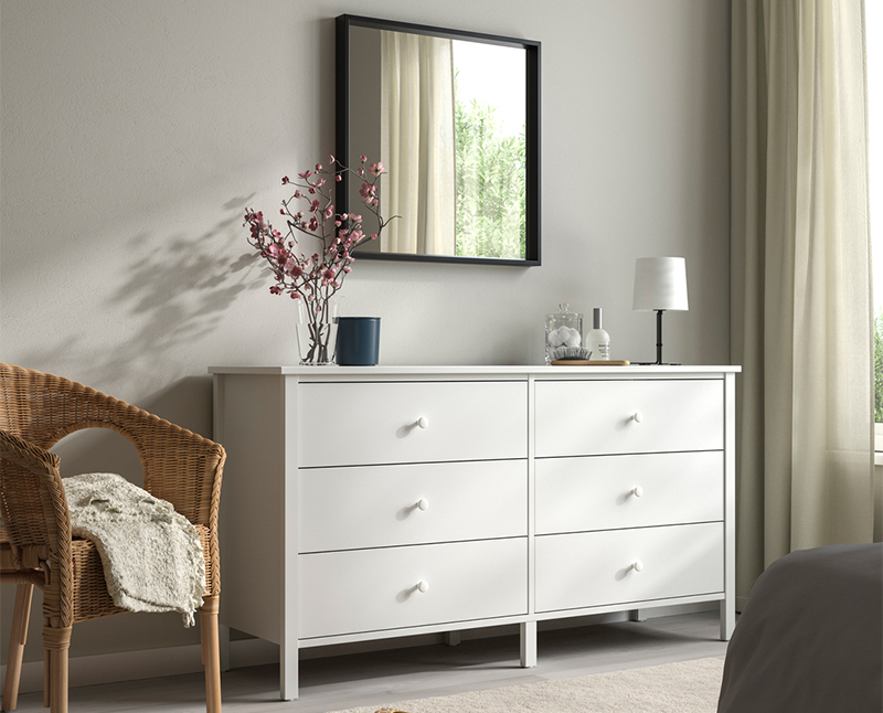 White six-drawer dresser with a square mirror, flower vase, and wicker chair.