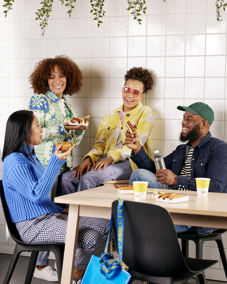A group of four friends laughing and sharing hot dogs and drinks around a table indoors.