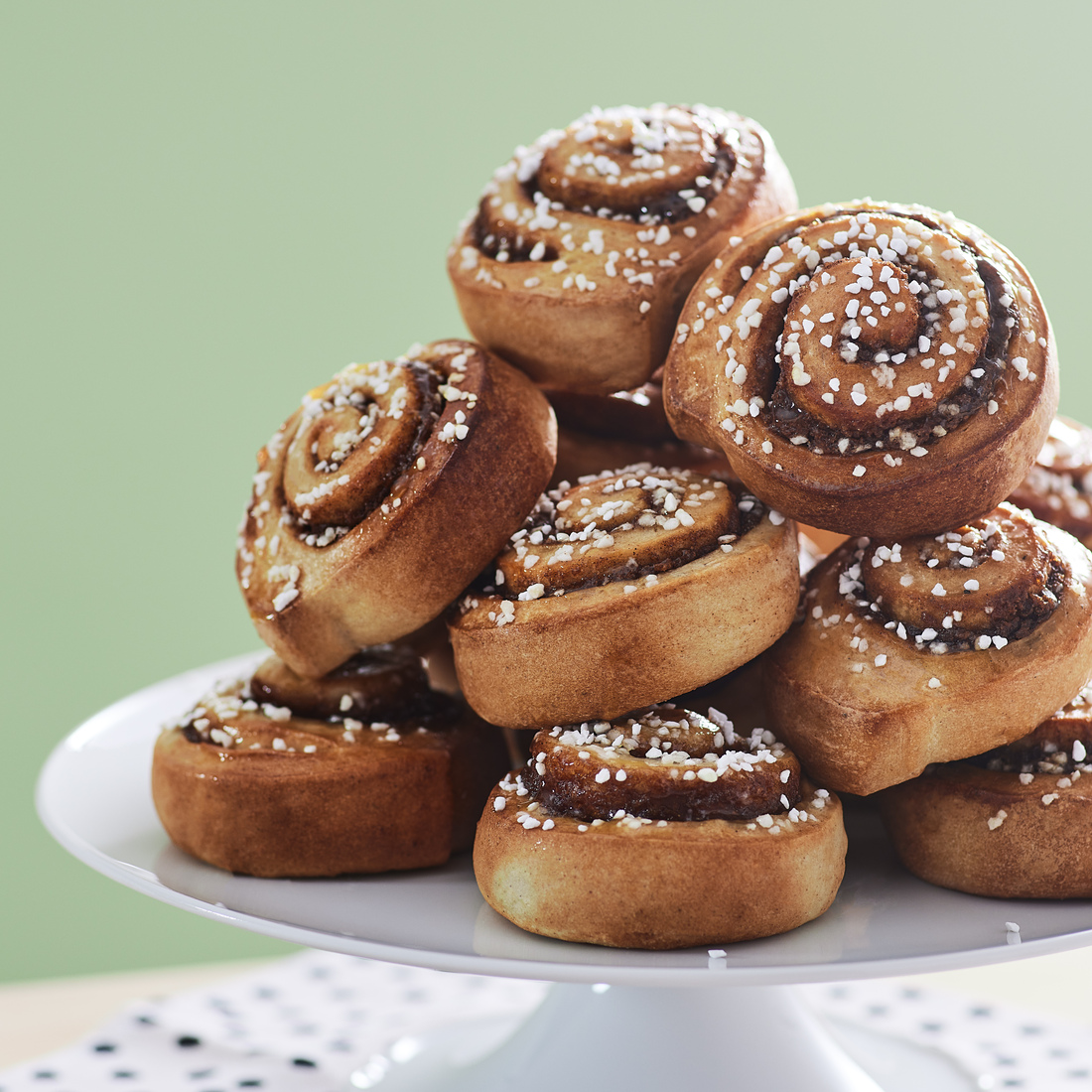A stack of freshly baked cinnamon buns sprinkled with pearl sugar, arranged on a white cake stand.