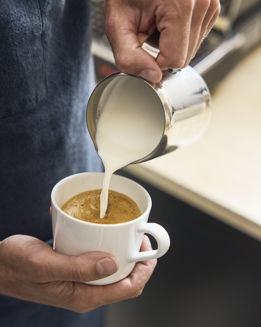 A person pouring steamed milk from a metal pitcher into a white cup of coffee to create latte art.