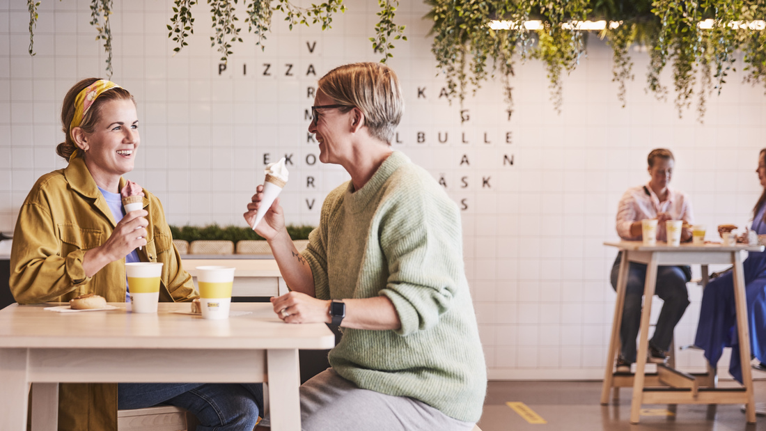 Two women smiling and chatting while eating ice cream cones in a cafe setting.